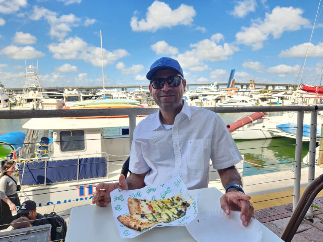 The photo shows a man, Rohit Godhani, sitting at an outdoor table by a marina.